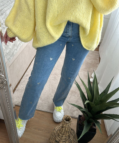 Frau in gelbem Pullover und Jeans mit Herzmuster, weiße Sneaker, Zimmerpflanze im Vordergrund.