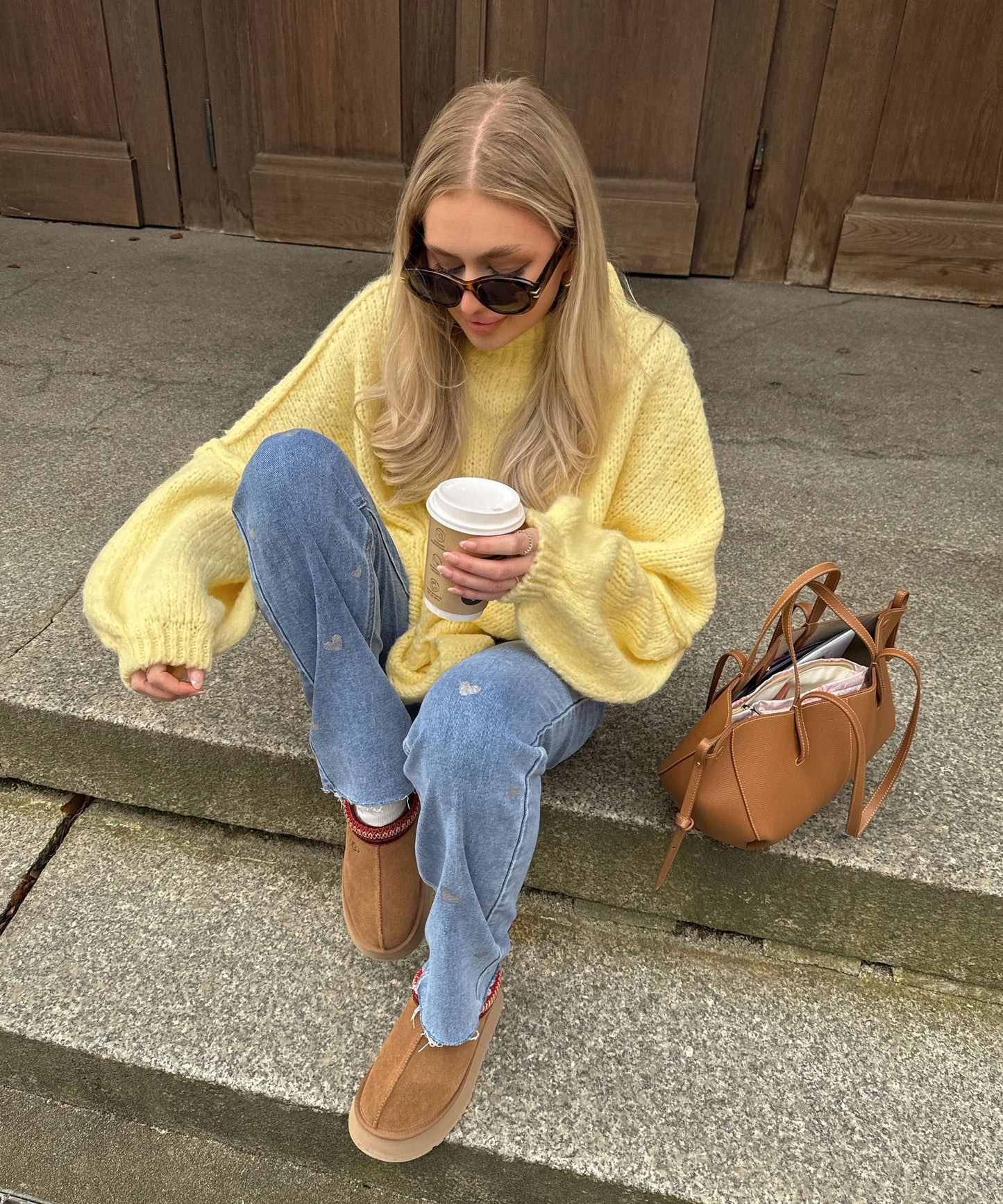Frau in gelbem Strickpullover, blauer Jeans, braunen Stiefeln, mit Sonnenbrille und Kaffeebecher.