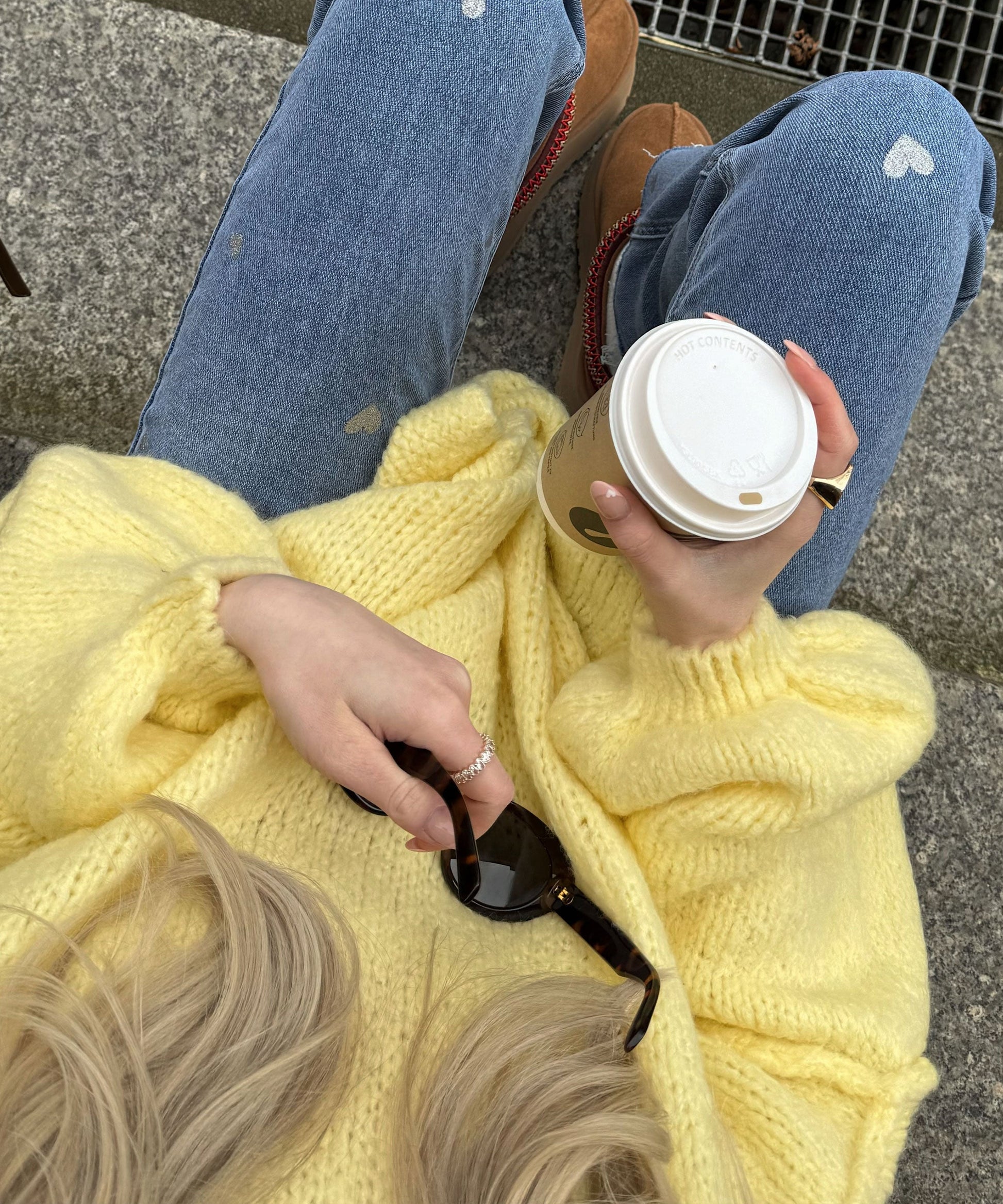 Frau in gelbem Strickpullover hält Kaffeebecher, trägt Jeans mit Herzmuster und Sonnenbrille.