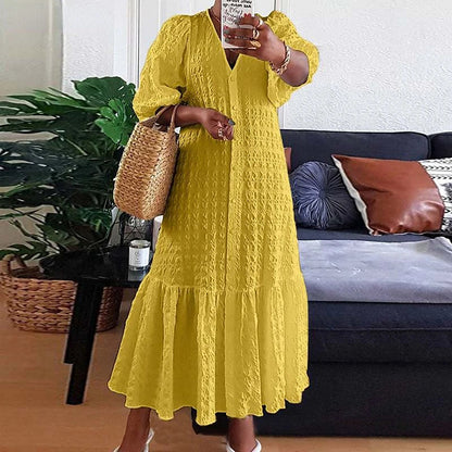 Frau in gelbem Sommerkleid mit Strohtasche vor Sofa und Pflanzen im Wohnzimmer.