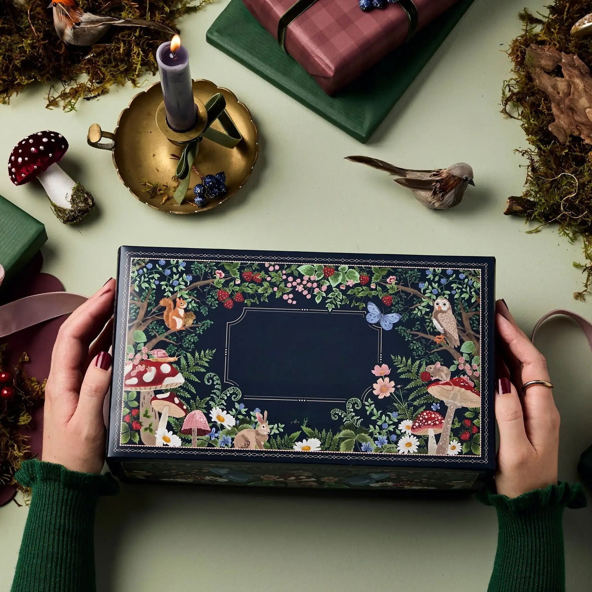 Hände halten dekorative Geschenkbox mit Waldmotiven, Pilzen und Tieren auf grünem Hintergrund.