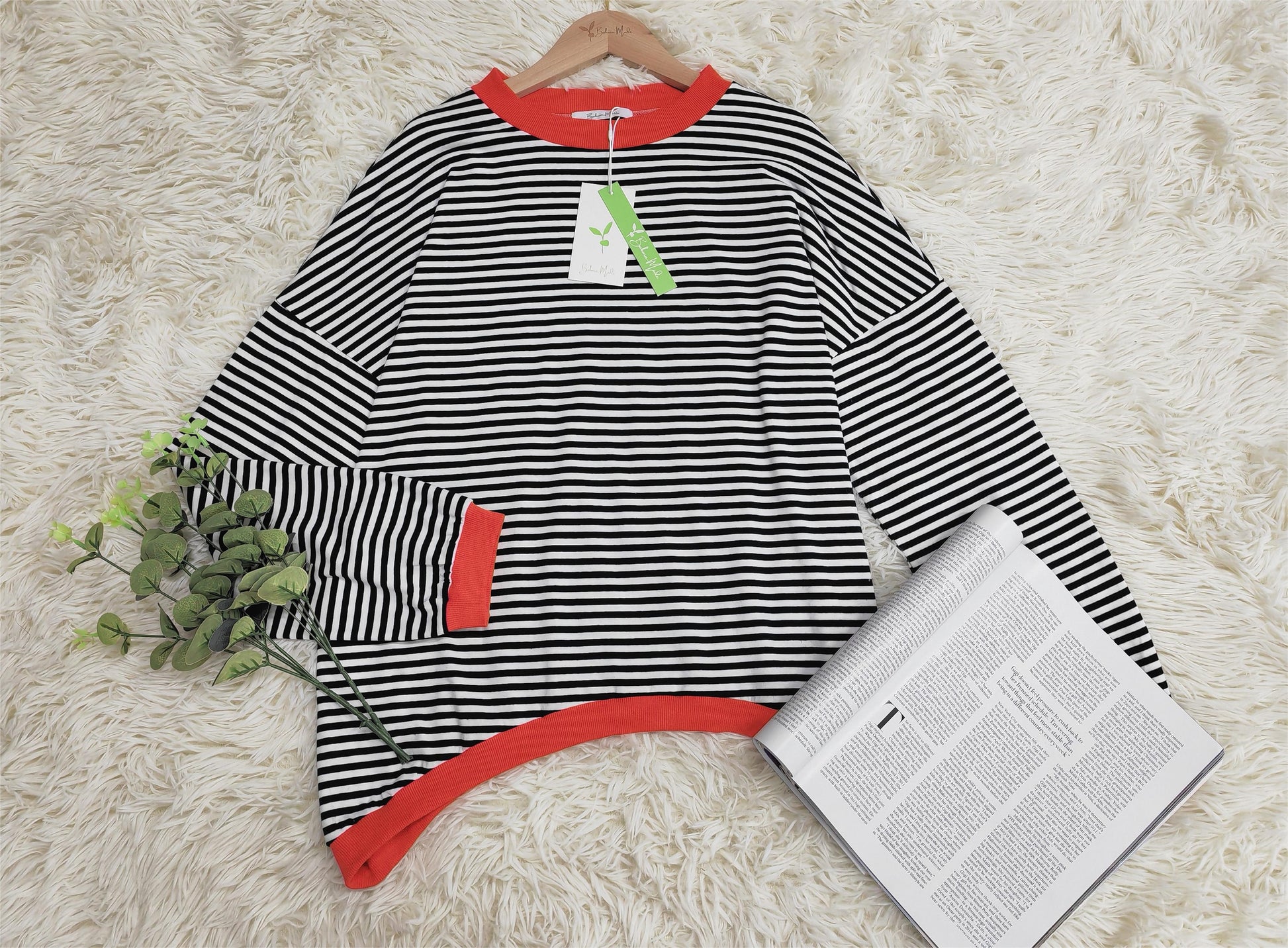 Schwarz-weiß gestreifter Pullover mit roten Bündchen auf Teppich, neben Zeitung und Pflanze.
