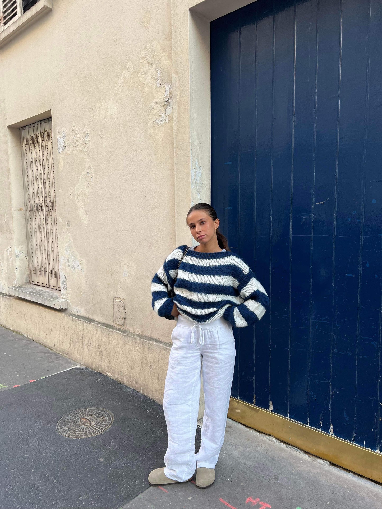 Frau in gestreiftem Pullover und weißer Hose vor blauer Tür, lässige Mode, Streetstyle.