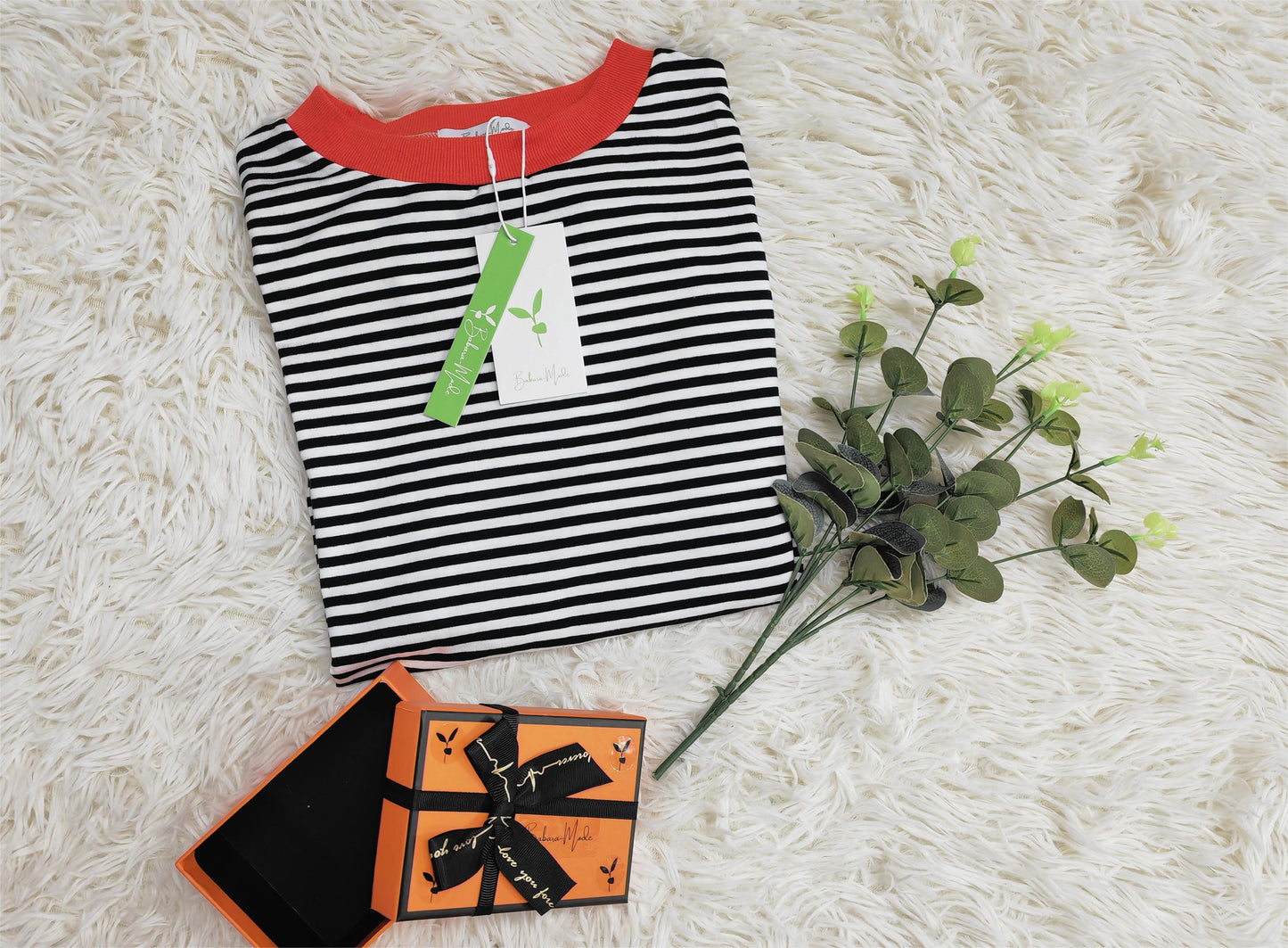 Gestreiftes T-Shirt mit rotem Kragen auf Teppich, Geschenkbox und grüner Zweig daneben.