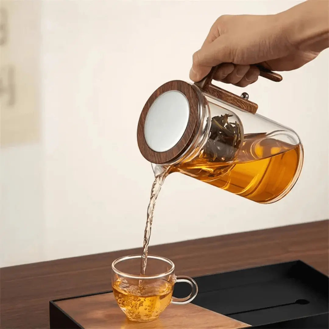 Hand gießt Tee aus Glas-Teekanne mit Holzgriff in Tasse, modernes Design, stilvoll, funktional.