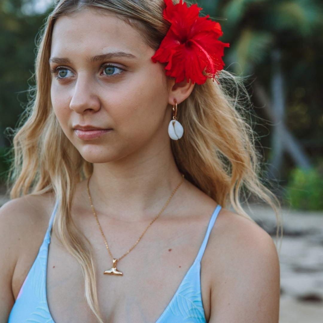 Frau mit goldener Walflossen-Halskette, roter Blume im Haar, Strandhintergrund.