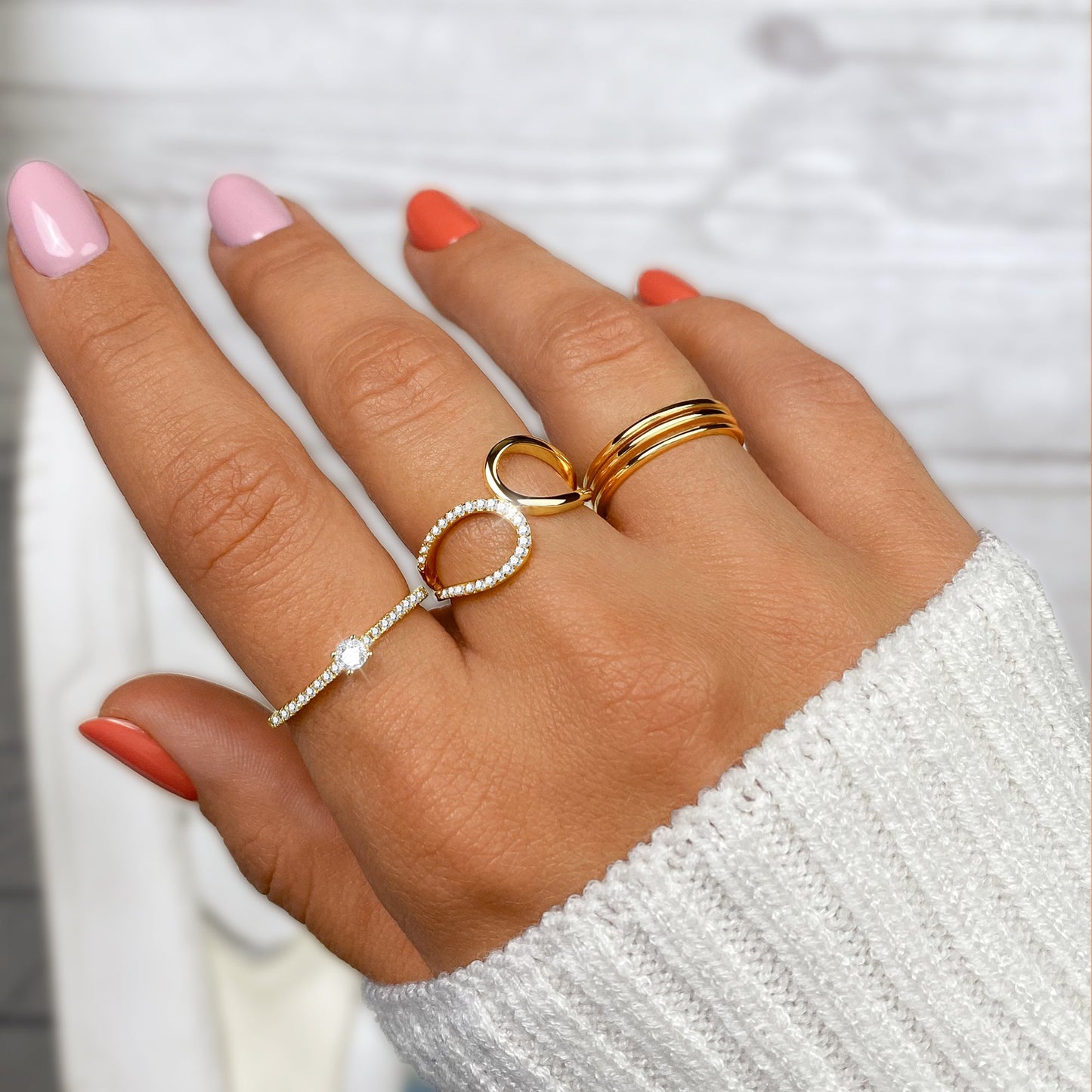Damenhand mit goldenen und silbernen Ringen, rosa und orange Nägel, weißer Pullover.