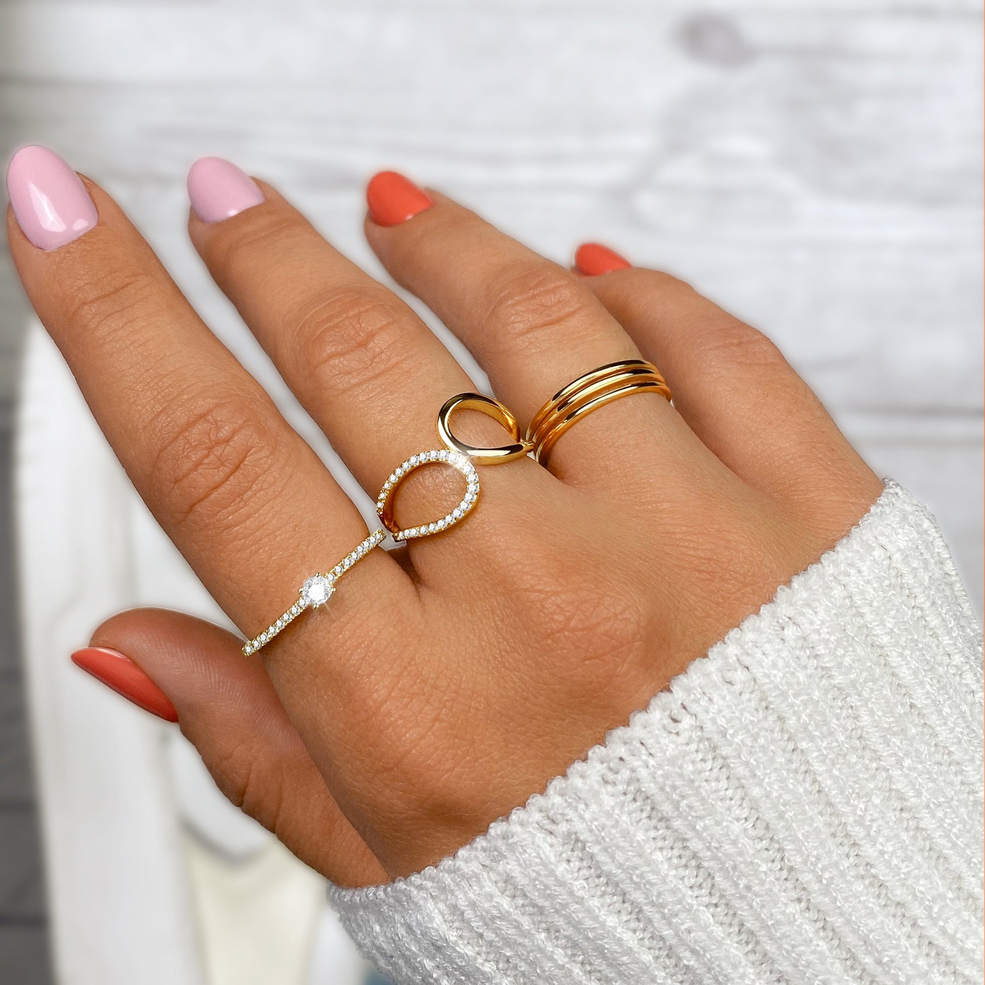Damenhand mit goldenen und silbernen Ringen, rosa und orange Nägel, weißer Pullover.