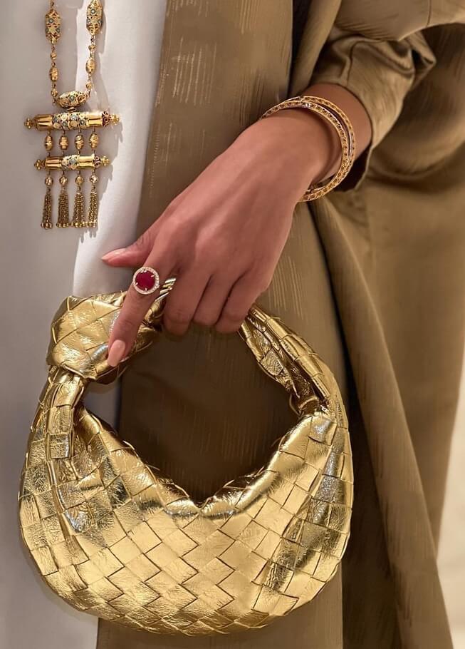 Frau mit goldener, geflochtener Handtasche, Schmuck und braunem Kleid.