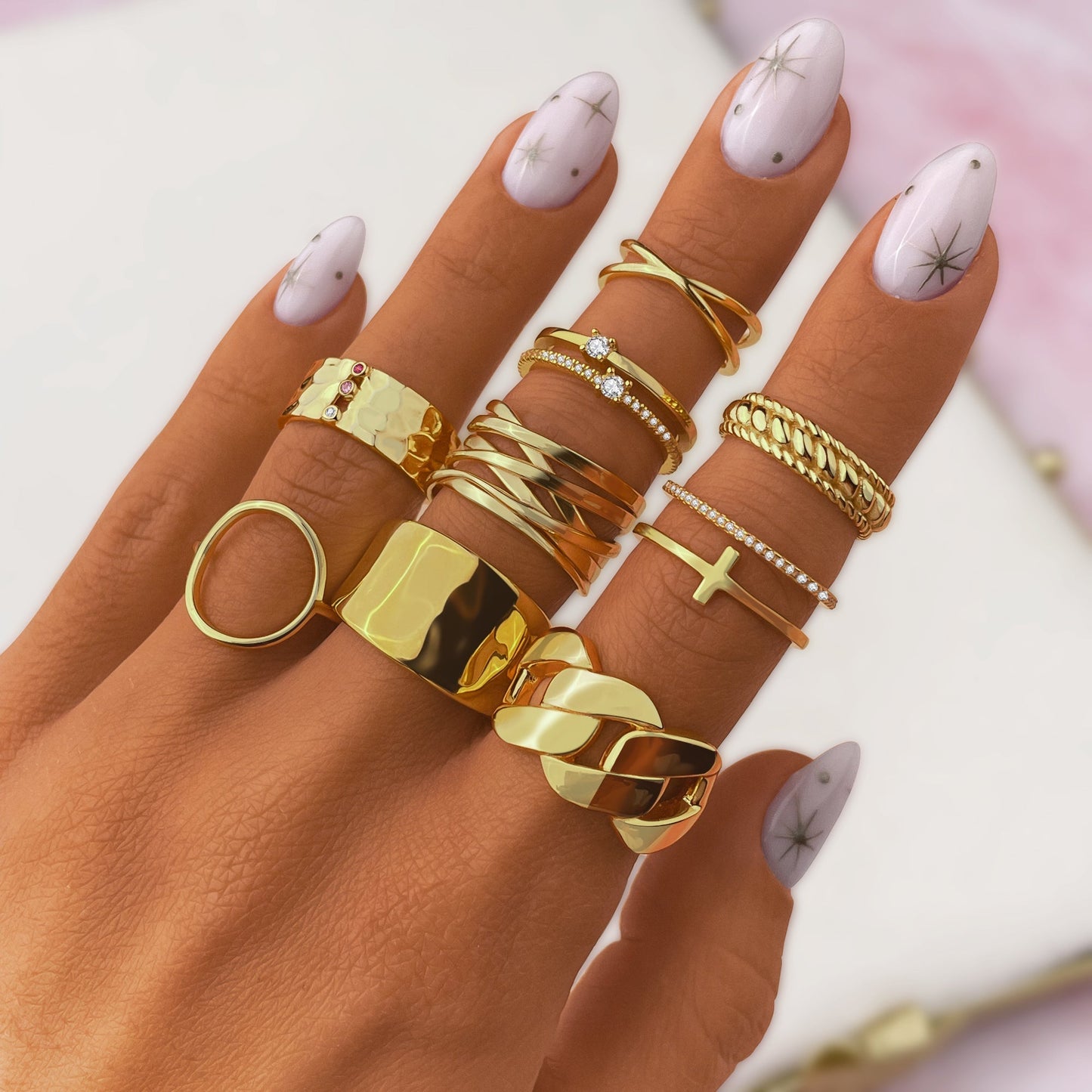 Hand mit goldenen Ringen, verschiedene Designs, auf pastellfarbenem Hintergrund, Nageldesign mit Sternen.