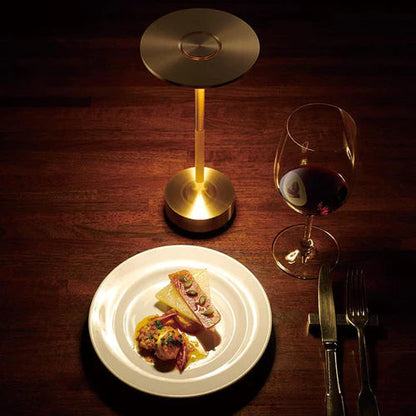Goldene Tischlampe aus Metall neben Teller mit Gourmet-Essen und Weinglas auf Holztisch.