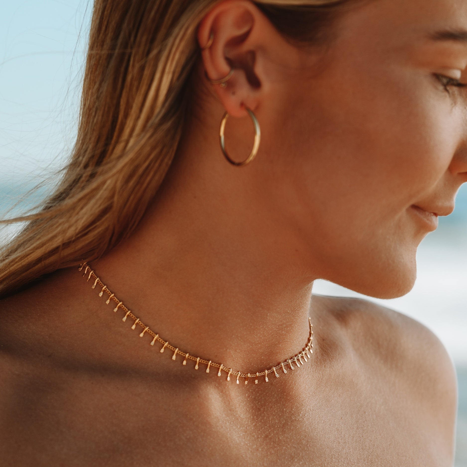 Frau mit goldener Choker-Halskette und Ohrring am Strand, modischer Schmuck.