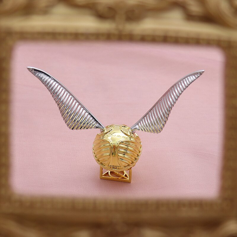 Goldener Schnatz Replik mit silbernen Flügeln, Harry Potter Sammlerstück, auf rosa Hintergrund.