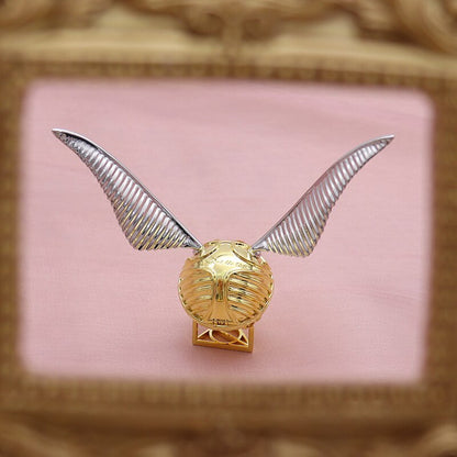 Goldener Schnatz Replik mit silbernen Flügeln, Harry Potter Sammlerstück, auf rosa Hintergrund.