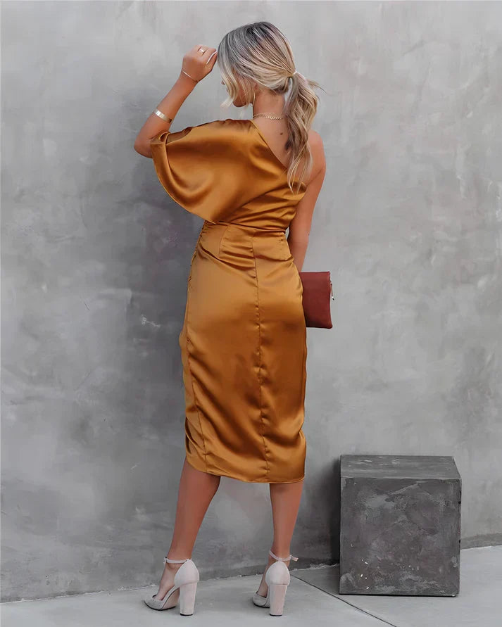 Frau in goldfarbenem Satin-Kleid mit asymmetrischem Schnitt, steht vor grauer Wand.