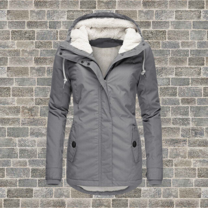 Graue Damen-Winterjacke mit Kapuze, Fleece-Futter, Reißverschluss, vor Ziegelwand.