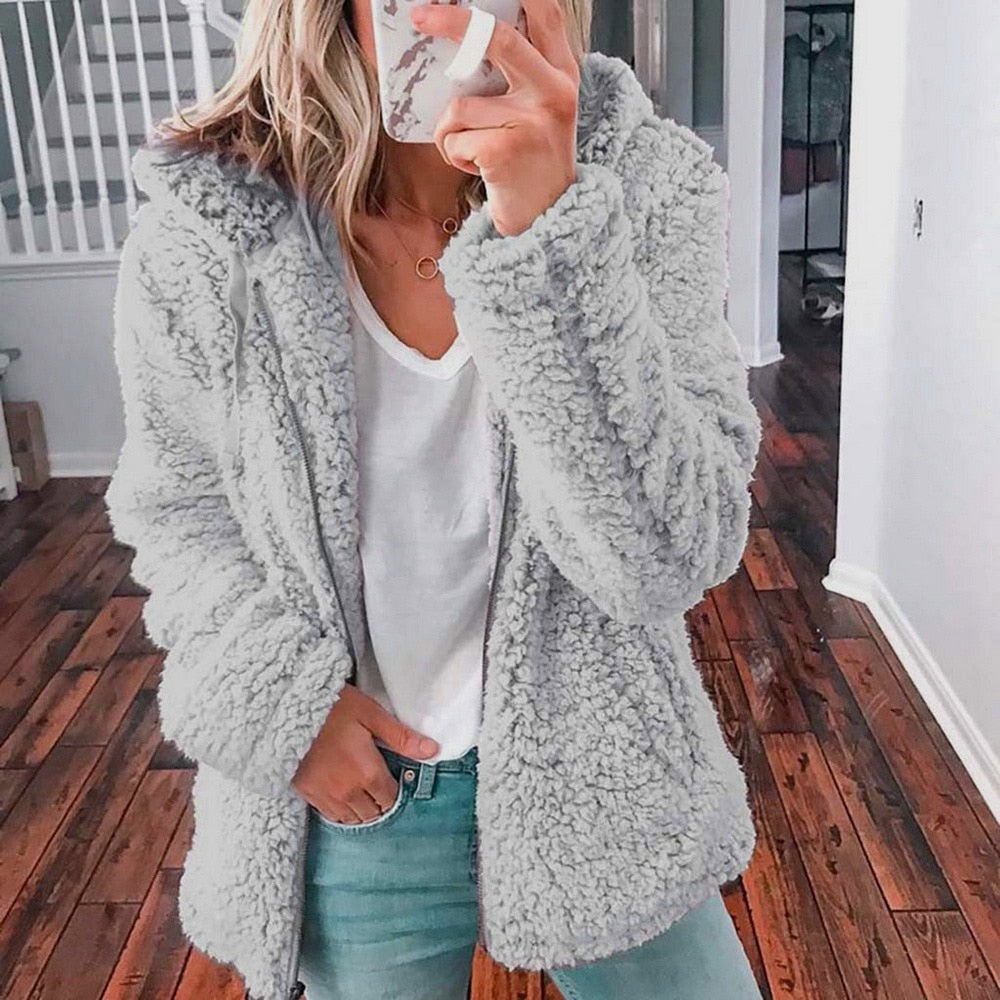 Frau in grauer Teddyjacke, weißem T-Shirt und Jeans, Selfie im Innenraum, Holzboden.