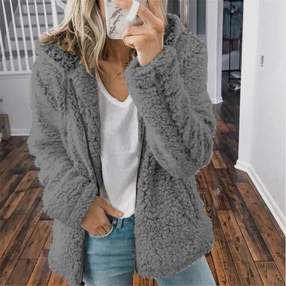 Frau in grauer Teddyjacke, weißem T-Shirt und Jeans, Smartphone in der Hand, Innenraum.