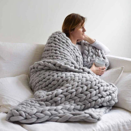 Frau mit grauer, grob gestrickter Decke auf Sofa, hält Tasse; gemütliche Wohntextilien.