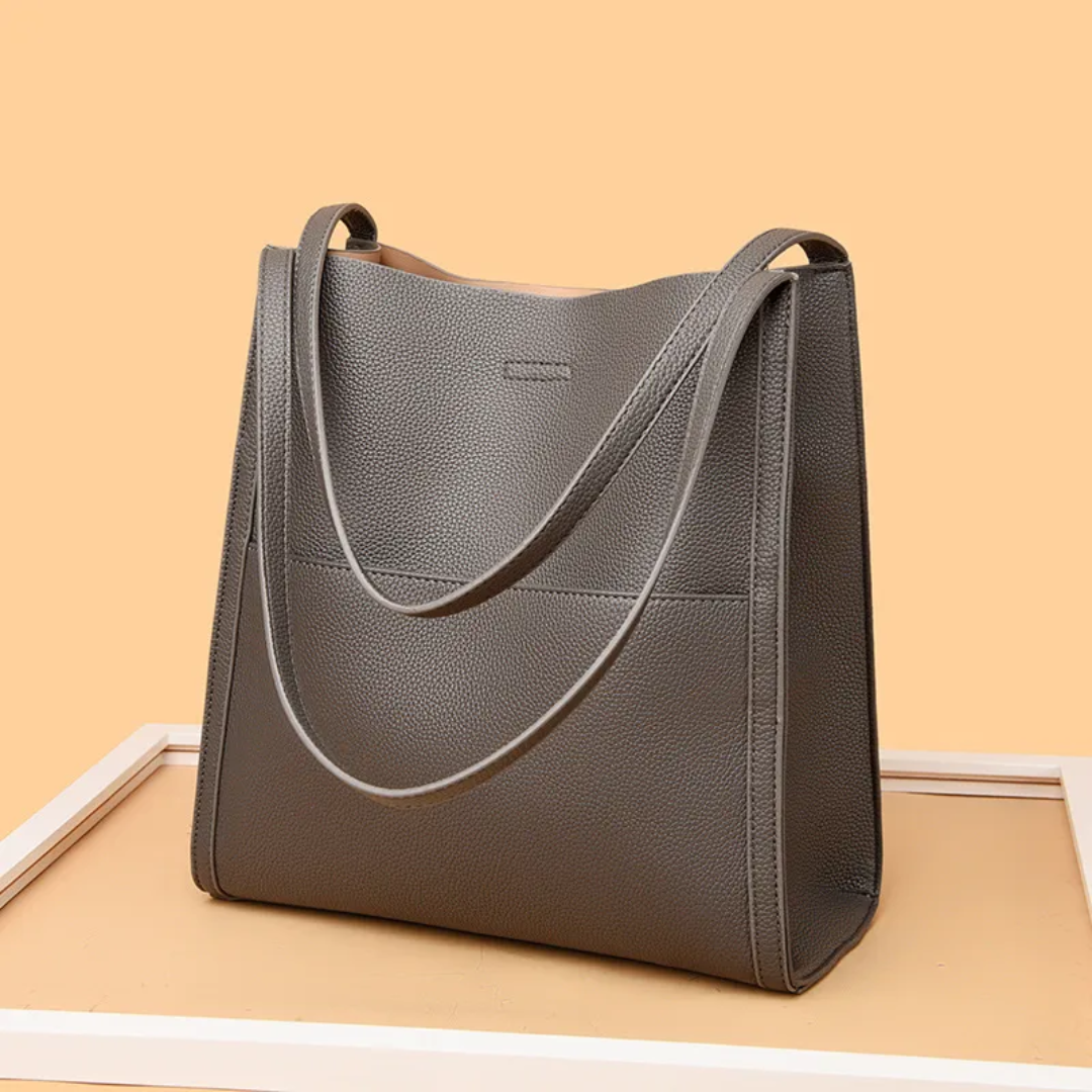 Graue Ledertasche mit langen Henkeln auf beigem Hintergrund, modisch und elegant.