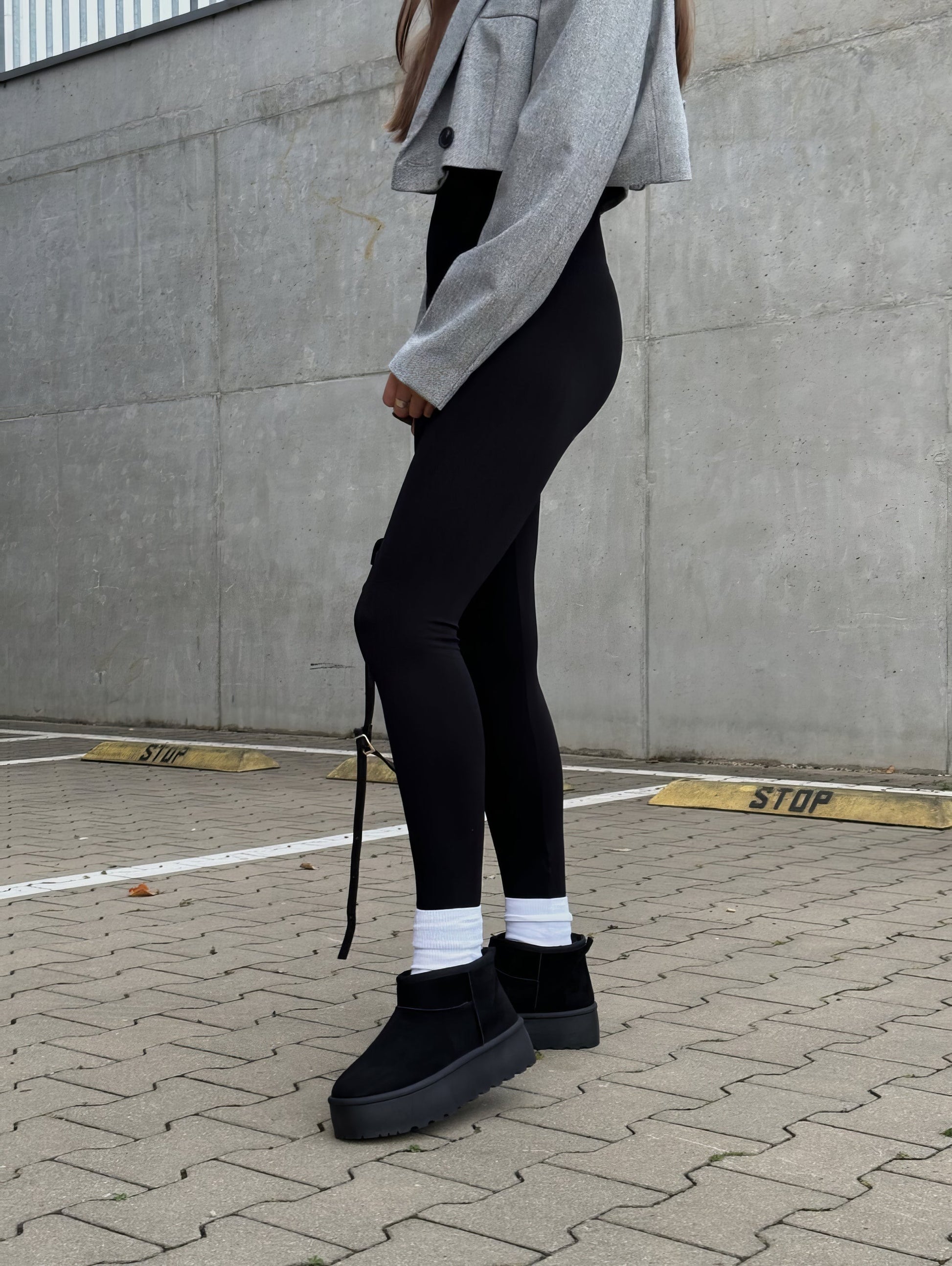 Frau in grauer Jacke, schwarzer Leggings und schwarzen Stiefeln auf gepflastertem Parkplatz.