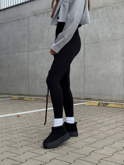 Frau in grauer Jacke, schwarzer Leggings und schwarzen Stiefeln auf gepflastertem Parkplatz.