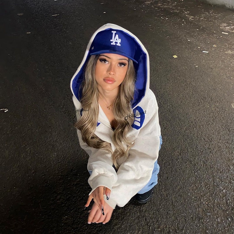 Frau in weißem Hoodie und blauer LA-Cap, kniend auf Asphalt.