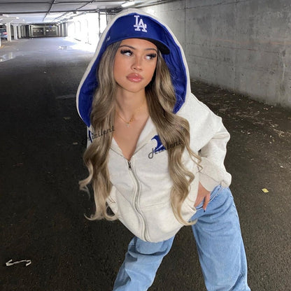 Frau in grauem Hoodie und blauer LA-Cap, lässiger Streetwear-Look, urbaner Hintergrund.
