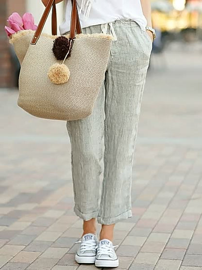 Frau mit Leinenhose, Strohtasche und weißen Sneakers auf gepflastertem Gehweg.