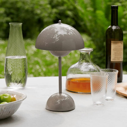 Graue Tischlampe aus Metall auf Gartentisch, umgeben von Karaffen und Gläsern im Freien.