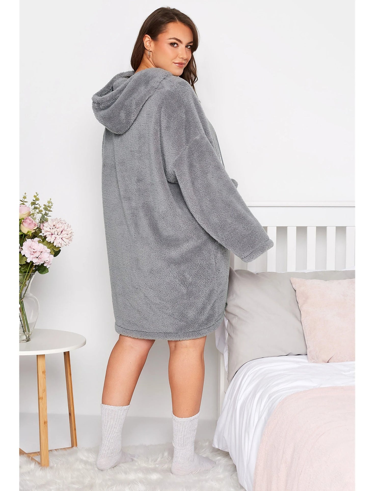 Frau in grauem Fleece-Bademantel mit Kapuze, steht neben Bett, gemütliche Wintermode.