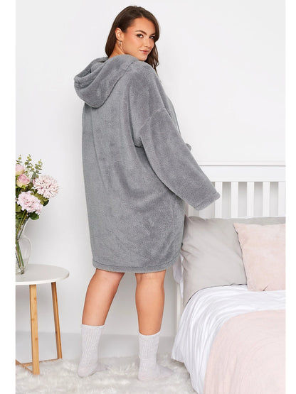 Frau in grauem Fleece-Bademantel mit Kapuze, steht neben Bett, gemütliche Wintermode.