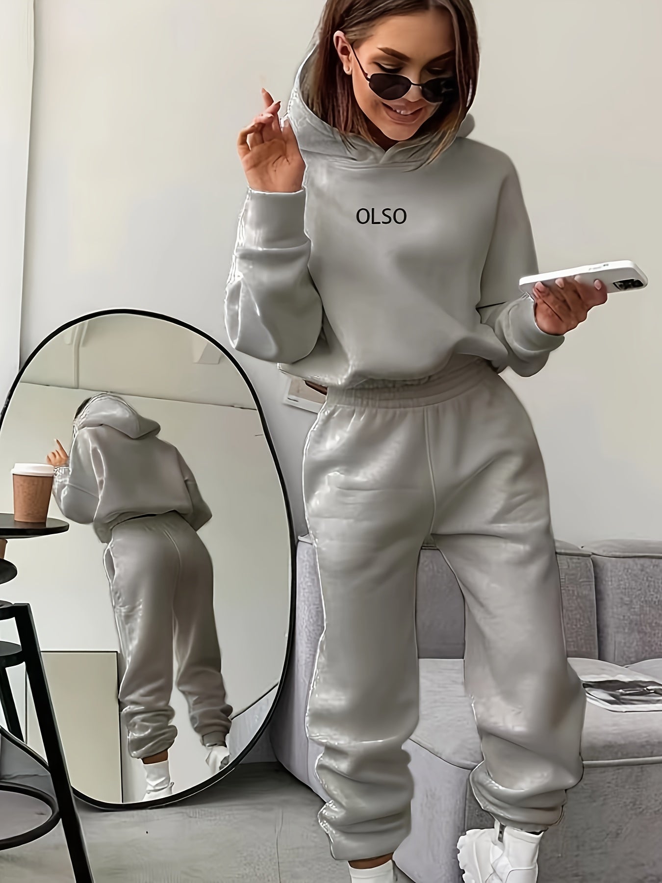 Frau in grauem Leder-Trainingsanzug mit Smartphone und Kaffeetasse, Spiegel im Hintergrund.