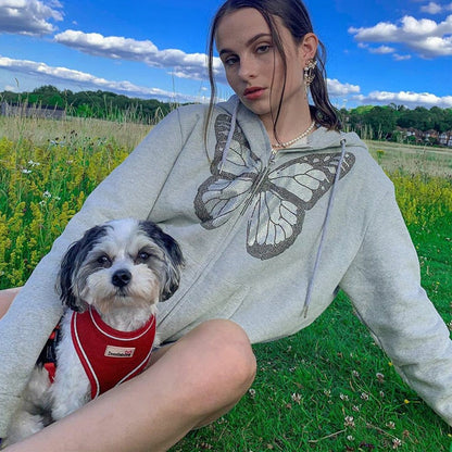 Frau in grauem Schmetterlings-Hoodie mit kleinem Hund im roten Geschirr auf Wiese.