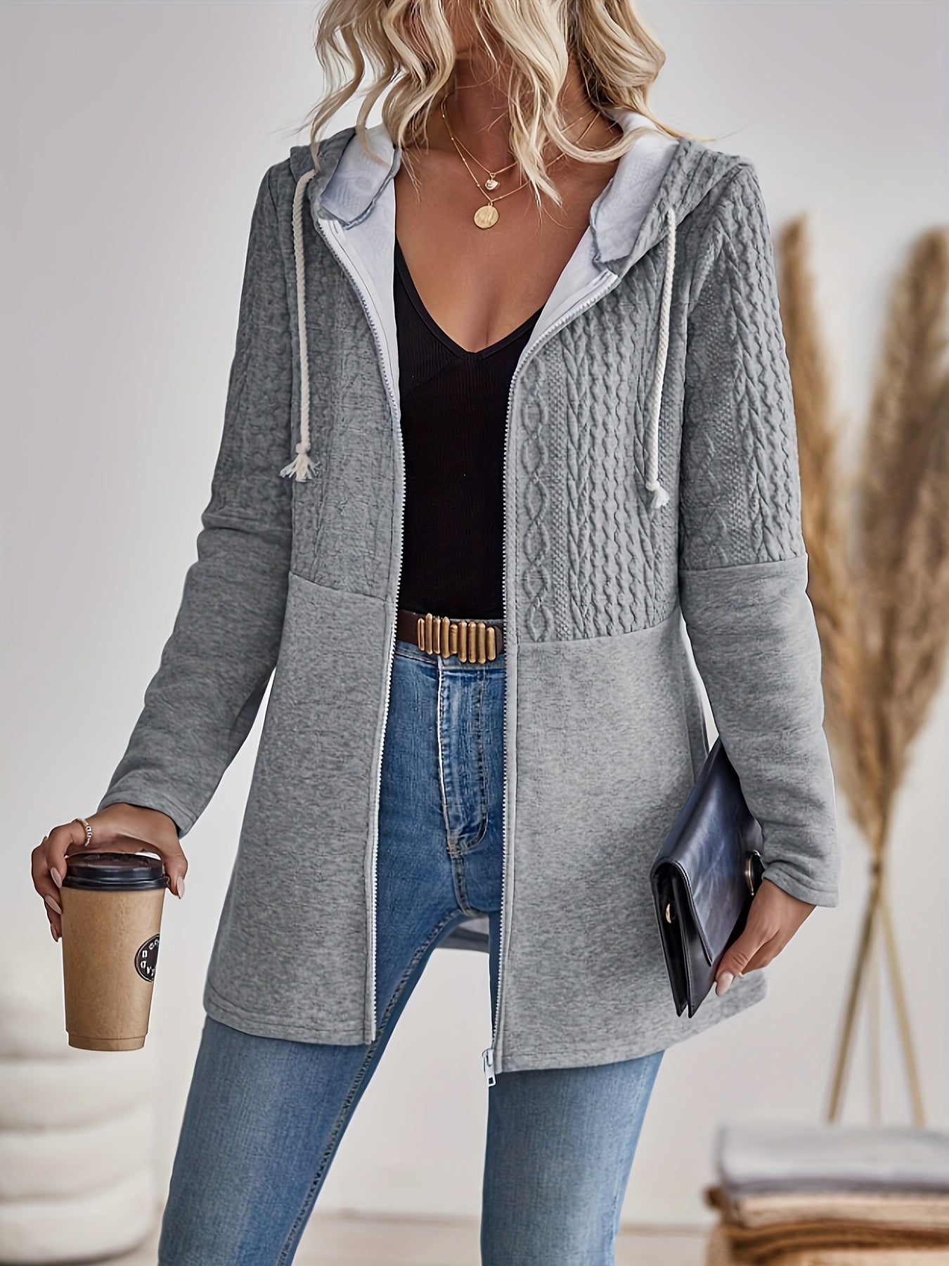 Frau in grauem Strickmantel mit Kapuze, hält Kaffeebecher und schwarze Clutch, modisch, lässig.