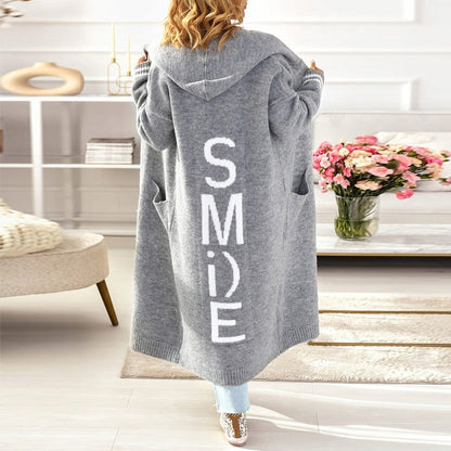 Frau in grauem Kapuzen-Cardigan mit "SMILE"-Aufdruck, Wohnzimmer, Blumenstrauß, gemütlich.