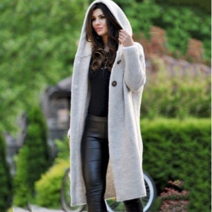Frau in grauem Strickmantel mit Kapuze, schwarze Lederhose, draußen im Grünen.