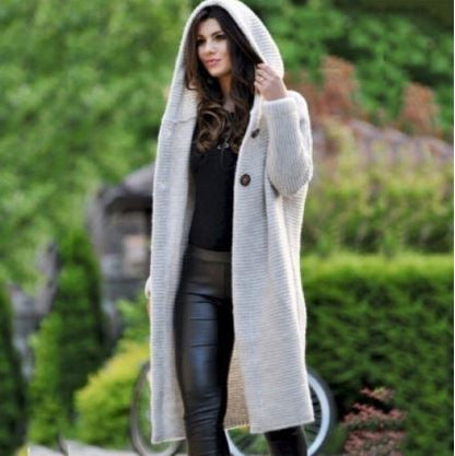 Frau in grauem Strickmantel mit Kapuze, schwarze Lederhose, draußen im Grünen.