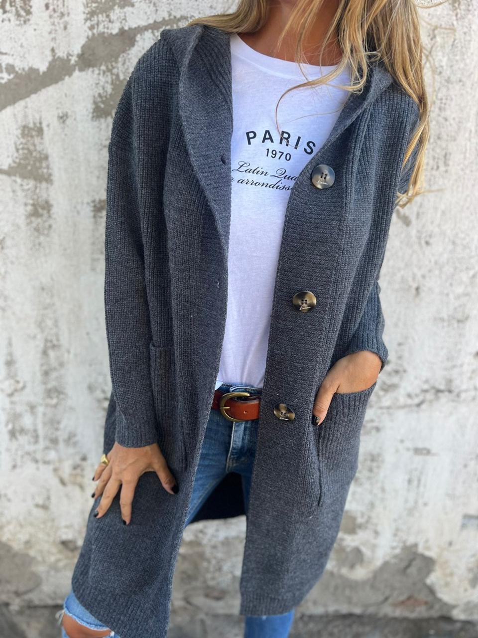 Frau in grauem Strickmantel mit Knöpfen, weißem T-Shirt und Jeans vor Betonwand.