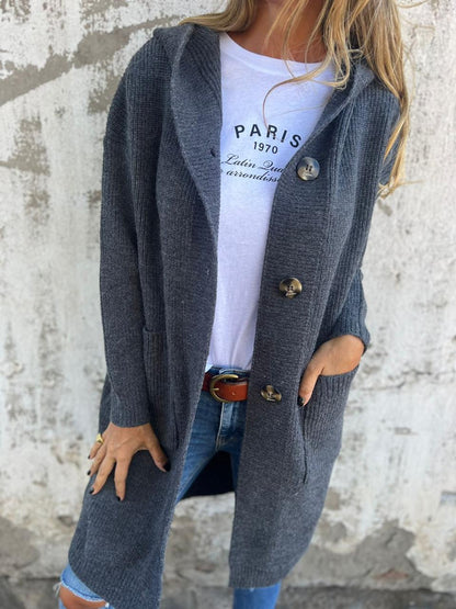 Frau in grauem Strickmantel mit Knöpfen, weißem T-Shirt und Jeans vor Betonwand.