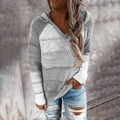 Frau in grauem Strickpullover mit Kapuze und zerrissenen Jeans vor Betonwand.