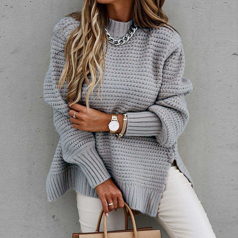 Frau in grauem Strickpullover, Kettenhalskette, weiße Hose, Armbanduhr, modisch, elegant.