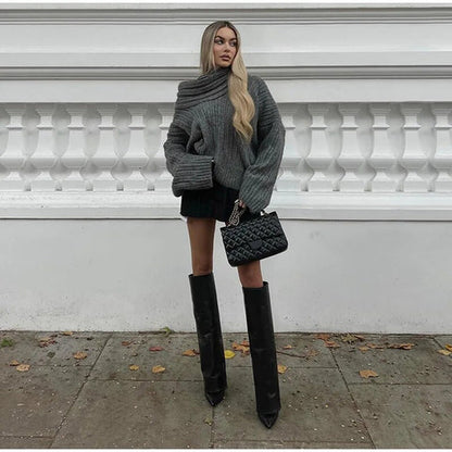 Frau in grauem Strickpullover mit einer Schulter, schwarze Stiefel, Tasche, Herbstmode.