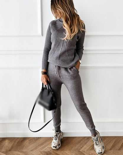 Frau in grauem Strickpullover und Jogginghose, trägt schwarze Handtasche und Sneakers.