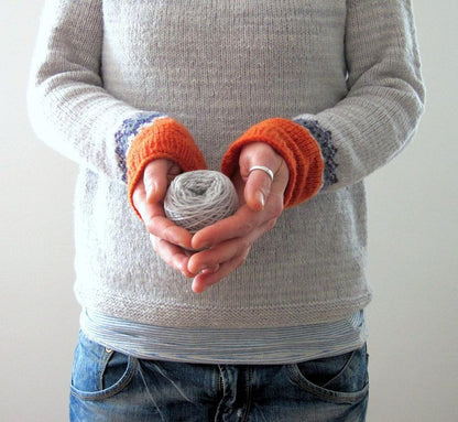 Person hält graues Wollknäuel, trägt grauen Pullover mit orangefarbenen Bündchen, blaue Jeans.