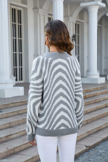 Frau in grauem Zickzack-Pullover, weiße Hose, vor klassischer Architektur.