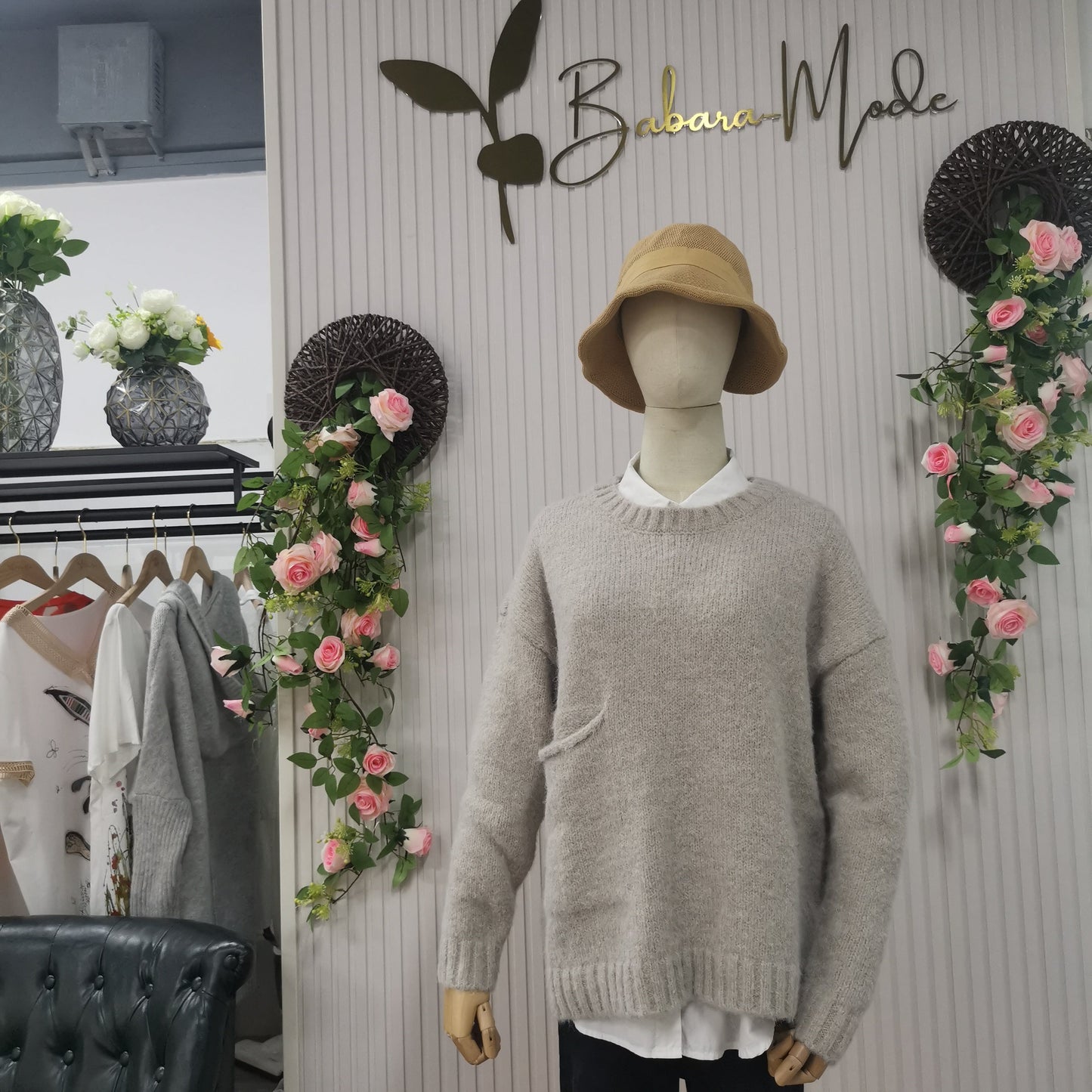Schaufensterpuppe mit grauem Pullover und beigem Hut, umgeben von Blumen, in Modegeschäft.