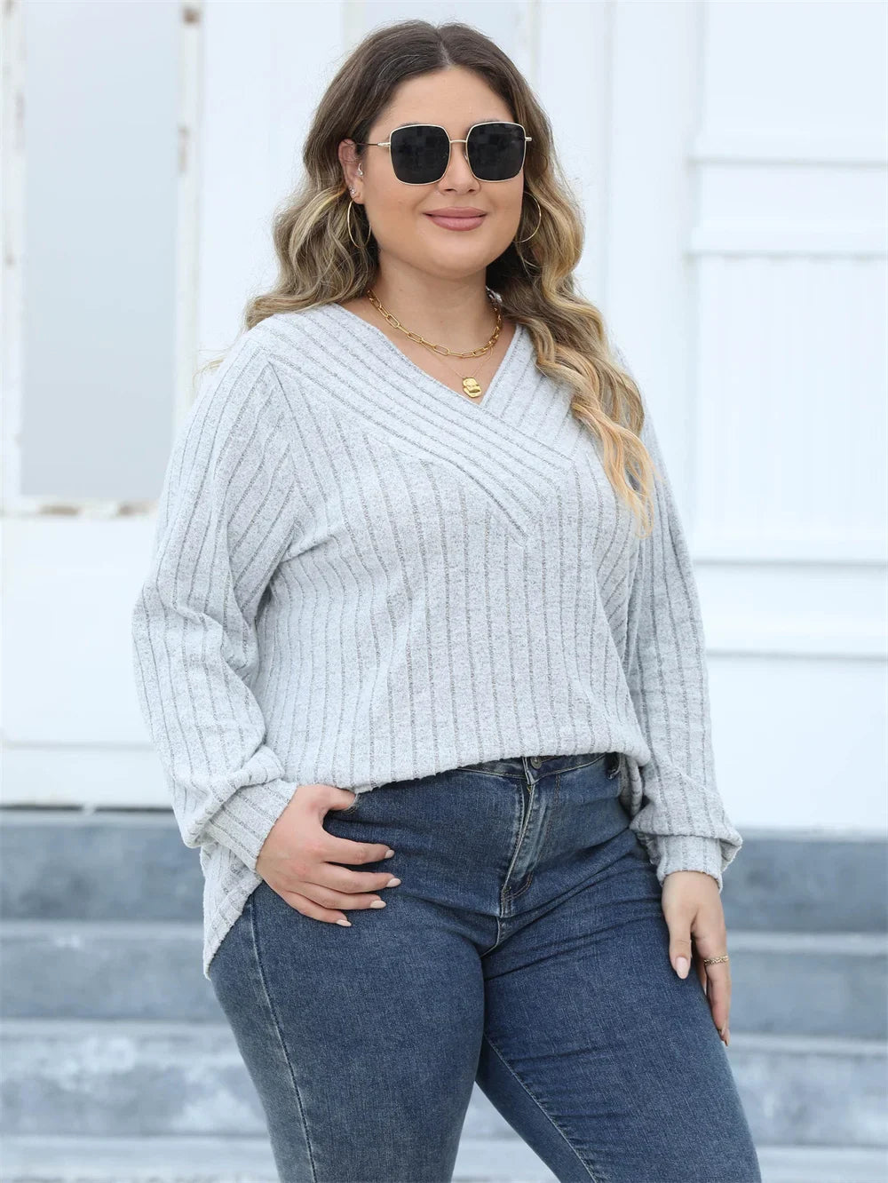 Frau in grauem Strickpullover und blauer Jeans, trägt Sonnenbrille, lächelt, steht draußen.