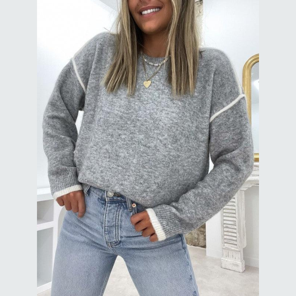 Frau trägt grauen Strickpullover, blaue Jeans, Herzanhänger; modisches Winteroutfit.
