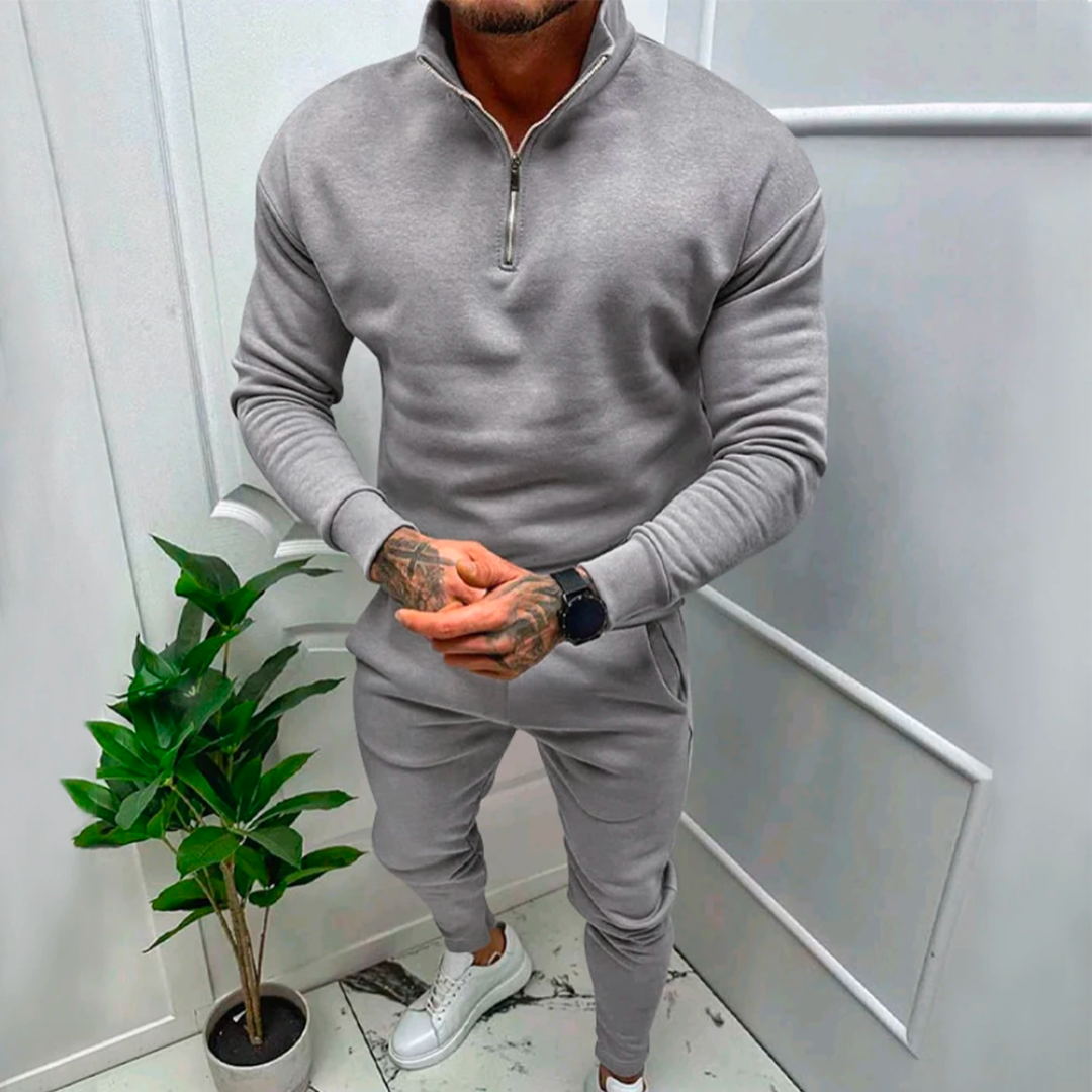 Mann in grauem Trainingsanzug mit Reißverschluss, weiße Sneaker, Zimmerpflanze, moderne Einrichtung.